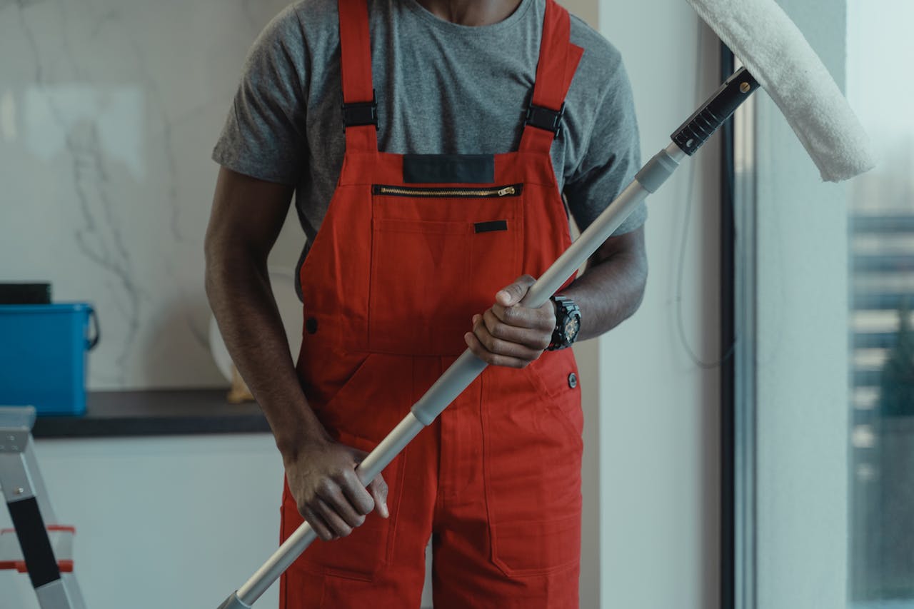 hero-img-01 A professional cleaner wearing red overalls is holding a mop while cleaning indoors.
